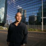 Elegant businessman posing confidently on a modern street in Brasília, with reflective skyscrapers in the background.
